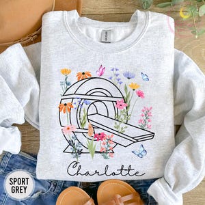 Könnte beinhalten: Sportgraues Sweatshirt mit einer schwarzen Strichzeichnung eines CT-Scanners, verziert mit bunten Blumen und Schmetterlingen. Der Name "Charlotte" steht darunter in Schreibschrift. Das Sweatshirt ist aus weichem Material.
