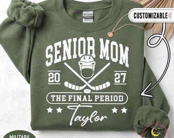 Op de afbeelding: Een olijfgroene sweater met ronde hals en witte opdruk met de tekst SENIOR MOM 20 27 THE FINAL PERIOD en de naam Taylor. Het ontwerp bevat een ijshockeyhelm, twee gekruiste ijshockeysticks, een puck en vier sterren.