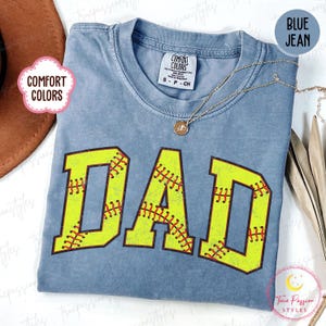 Op de afbeelding: Blauw Comfort Colors T-shirt met het woord "DAD" in grote, gele letters. De letters zijn ontworpen om op softballs te lijken, met rode stikseldetails. Een gouden ketting is over het shirt gedrapeerd.