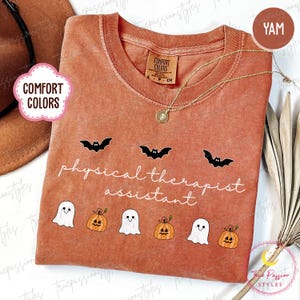 May include: A yam-colored Comfort Colors t-shirt with the text "physical therapist assistant" and Halloween-themed graphics of bats, ghosts, and pumpkins. A gold necklace with a pendant is draped on the shirt.