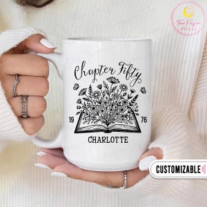 May include: A white ceramic mug featuring a black line art illustration of an open book with a bouquet of wildflowers growing from the pages. The text reads Chapter Fifty, 1976, and the name Charlotte. The design includes small butterflies.