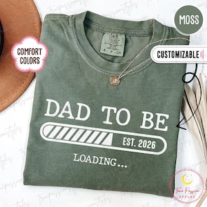 May include: Moss green t-shirt with the text "DAD TO BE" and "EST. 2026 LOADING..." in white. The shirt has a crew neck and short sleeves. A silver necklace with a small pendant is visible. The shirt is labeled "Comfort Colors".