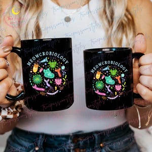 Puede incluir: Dos tazas de caf&eacute; negras con un dise&ntilde;o blanco. El dise&ntilde;o presenta un c&iacute;rculo con el texto "MEOWCROBIOLOGY" y gatos y bacterias de dibujos animados coloridos.