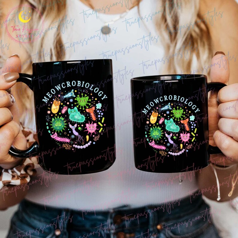 Puede incluir: Dos tazas de caf&eacute; negras con un dise&ntilde;o blanco. El dise&ntilde;o presenta un c&iacute;rculo con el texto "MEOWCROBIOLOGY" y gatos y bacterias de dibujos animados coloridos.
