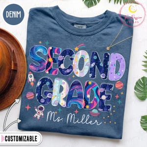 Puede incluir: Una camiseta azul denim con el texto SECOND GRADE en letras grandes con temática espacial, rellenas de patrones de galaxias, planetas y estrellas. Debajo del texto, el nombre Ms. Miller está impreso en letra blanca, junto a gráficos espaciales.