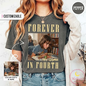 Pode incluir: Uma t-shirt cinzento-carvão de manga curta com uma impressão fotográfica retangular de uma criança a ler um livro. O texto "FOREVER IN FOURTH" está impresso a amarelo acima e abaixo da foto, com "Mrs. Sophia" escrito em cursivo.
