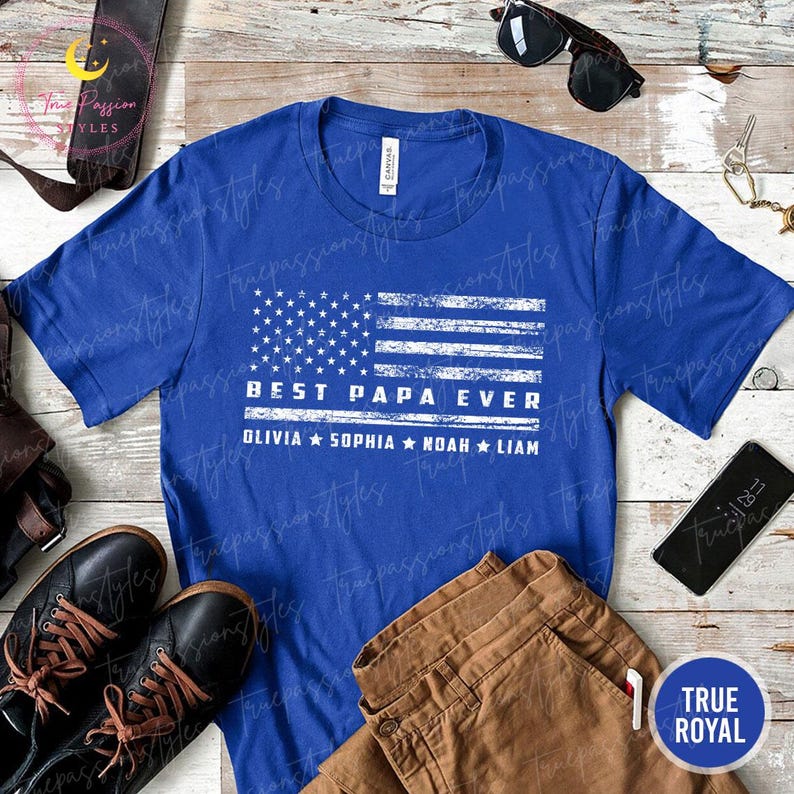 K&ouml;nnte beinhalten: Ein k&ouml;nigsblaues T-Shirt mit einem verblassten wei&szlig;en Design der amerikanischen Flagge. Der Text "BEST PAPA EVER" ist unter der Flagge aufgedruckt, darunter die Namen Olivia, Sophia, Noah und Liam. Das Shirt wird mit braunen Hosen und schwarzen Schuhen gezeigt.