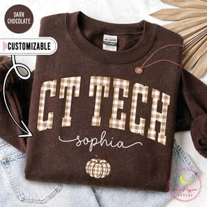 May include: A dark chocolate brown crewneck sweatshirt featuring the text CT TECH in a beige and white gingham pattern. Below the text, the name Sophia is written in white cursive script, followed by a small gingham patterned pumpkin graphic.