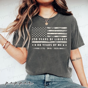 May include: A dark grey t-shirt with a distressed American flag graphic and the text "250 YEARS OF LIBERTY" and "80 YEARS OF ME", plus the years 1776-1946-2026. This is a casual wear item.