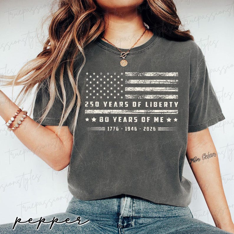 May include: A dark grey t-shirt with a distressed American flag graphic and the text "250 YEARS OF LIBERTY" and "80 YEARS OF ME", plus the years 1776-1946-2026. This is a casual wear item.