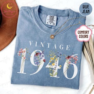 Op de afbeelding: Blauw spijkerbroekkleurig T-shirt met de witte woorden "VINTAGE 1946". De cijfers zijn versierd met kleurrijke bloemstukken. Een gouden ketting met een hanger ligt op het shirt. Het shirt heeft een Comfort Colors-label.