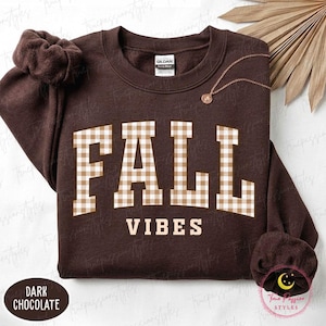May include: A dark chocolate brown crewneck sweatshirt featuring the text FALL in a beige and white gingham pattern, with the word VIBES printed in a solid beige font underneath. The garment includes a matching brown scrunchie and a gold necklace.