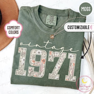 May include: A moss green t-shirt featuring the word "vintage" and the number "1971" in a floral pattern. The shirt has a crew neck and short sleeves. The text "Comfort Colors" is visible on the shirt.