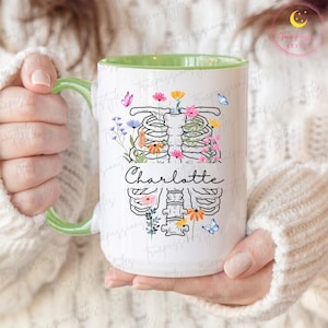 May include: White ceramic mug with a green handle and interior. The mug features a floral rib cage design with the name "Charlotte" in script. Colourful flowers and butterflies decorate the design, creating a unique and artistic look.