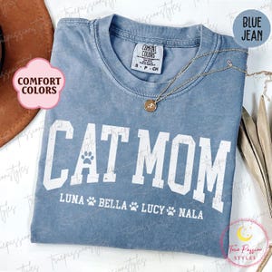 May include: A blue Comfort Colors t-shirt with the words "CAT MOM" in white, a paw print, and the names LUNA, BELLA, LUCY, and NALA. A gold necklace with a pendant is draped on the shirt. The shirt is a blue jean colour.