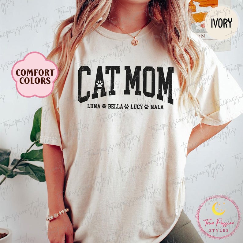 May include: Cream-coloured t-shirt featuring the text "CAT MOM" in bold, black lettering. Underneath are the names Luna, Bella, Lucy, and Nala. The shirt also has a paw print graphic. The words "Comfort Colors" are in a pink bubble.