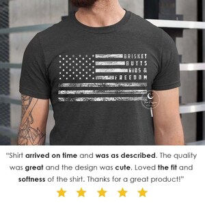 May include: A dark grey t-shirt with a distressed white American flag design. The text "BRISKET BUTTS RIBS & FREEDOM" is printed on the flag.