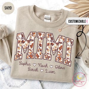 Könnte beinhalten: Ein sandfarbenes Sweatshirt mit Rundhalsausschnitt und dem Wort MIMI in großen Blockbuchstaben, die mit einem Blumenmuster in Orange, Braun und Weiß gefüllt sind. Unter dem Text sind die Namen Sophia, Noah, Olivia, Sarah und Liam aufgedruckt.