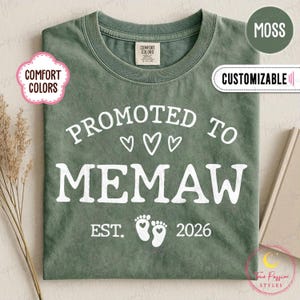 Könnte beinhalten: Ein moosgrünes T-Shirt mit dem weißen Schriftzug "PROMOTED TO MEMAW", drei Herzen, Babyfüßen und dem Jahr "2026". Das Shirt hat einen Rundhalsausschnitt und kurze Ärmel. Die Wörter "COMFORT COLORS" und "CUSTOMIZABLE" sind ebenfalls sichtbar.