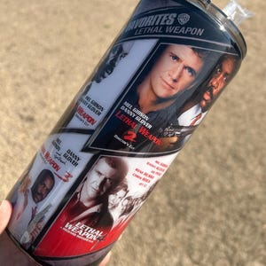 May include: A black and red stainless steel tumbler with a collage of images from the Lethal Weapon movie franchise. The tumbler features the text "Favorites Lethal Weapon" and the movie titles "Lethal Weapon 2", "Lethal Weapon 3", and "Lethal Weapon 4".