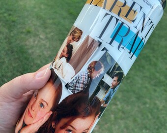 May include: A white tumbler featuring images from the movie "The Parent Trap." The tumbler has a black lid and a straw. The text "Walt Disney Pictures presents" and "The Parent Trap" are visible on the tumbler.