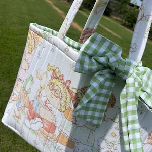 May include: A quilted fabric tote bag featuring a vintage-style illustration of a child with blonde yarn hair reading a book, accented with a green and white gingham bow tied to the handle. The bag has vertical stitching and floral patterns.