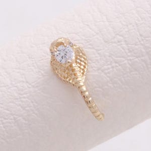 May include: A gold-colored ring in the shape of a cobra, with a clear, round gemstone set in the cobra's head. The ring is displayed on a white surface.
