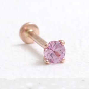 May include: A single, rose gold-colored stud earring with a light pink, round gemstone. The earring has a flat back and a post for piercing. The gemstone is held in place by four small prongs.