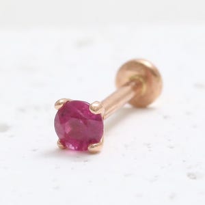 Może przedstawiać: Pojedynczy kolczyk w kolorze różowego złota z okrągłym, żywym, magentowym kamieniem szlachetnym. Widoczny jest sztyft i zapięcie kolczyka. Kamień jest osadzony w czterech pazurach, na białej, teksturowanej powierzchni.