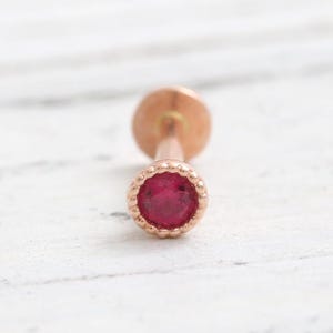 May include: A rose gold lip piercing stud with a round, bright red gemstone. The stud has a decorative, beaded bezel setting around the gemstone. The post is smooth and cylindrical.