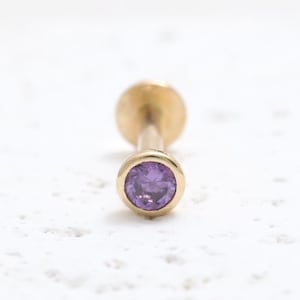 May include: A gold stud earring featuring a round, bezel-set purple gemstone. The earring post is straight with a flat, circular back. The gemstone is a vibrant purple, set within the gold metal setting.