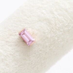 May include: A rose gold stud earring featuring a rectangular, light pink gemstone. The earring is set against a soft, white, textured background. The gemstone is approximately 0.6 cm long.