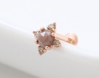 Könnte beinhalten: Ein Körperschmuckstück aus Roségold mit einem einzigen, runden, braunen Edelstein, der von kleineren, klaren Edelsteinen umgeben ist. Das Stück ist zum Tragen in einem Piercing gedacht.