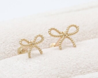 May include: A pair of gold-colored bow-shaped stud earrings. The bows have a twisted rope design and are set against a soft, off-white background. The earrings are small and delicate.