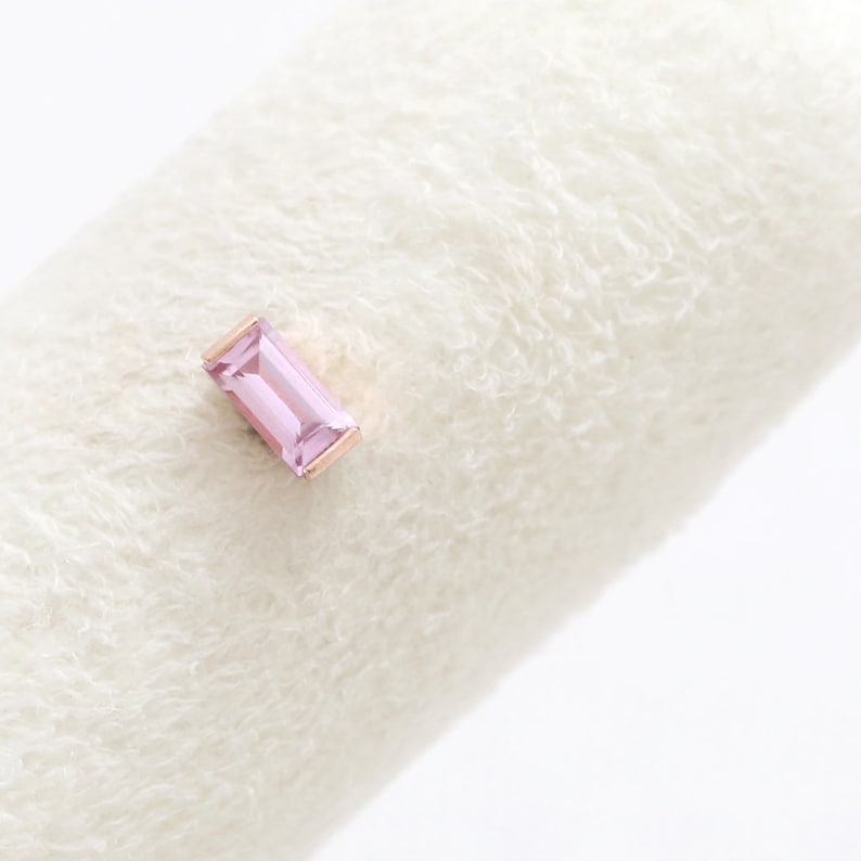May include: A rose gold stud earring featuring a rectangular, light pink gemstone. The earring is set against a soft, white, textured background. The gemstone is approximately 0.6 cm long.