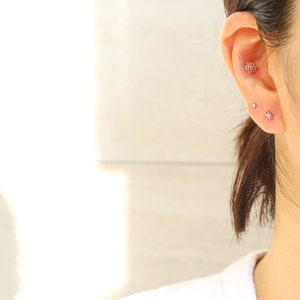 May include: Close-up of an ear with three earrings. The top earring is a rose-shaped stud. Below it is a small, round stud, and the third is a rectangular, pink stud. The ear is attached to a person wearing a white shirt.