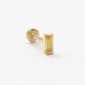 May include: A gold stud earring featuring a rectangular, yellow gemstone. The earring has a flat back and a post for piercing. The gemstone is a bright, clear yellow colour.