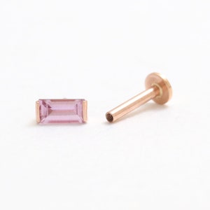May include: A rose gold labret stud and a matching baguette-cut pink gemstone. The stud has a flat disc backing and a threaded post. The gemstone is rectangular with a light pink hue, set in a rose gold bezel.