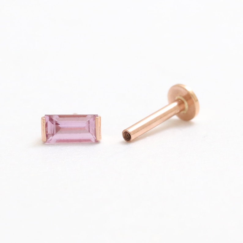 May include: A rose gold labret stud and a matching baguette-cut pink gemstone. The stud has a flat disc backing and a threaded post. The gemstone is rectangular with a light pink hue, set in a rose gold bezel.