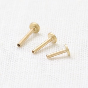 以下が含まれることがあります： 平らな円形のトップとストレートのポストを持つ、ゴールドカラーのボディジュエリーが3つ。ジュエリーは白い表面に並べて置かれています。