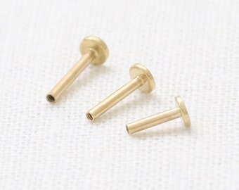 May include: Three gold-coloured body jewellery pieces, each with a flat circular top and a straight post. The jewellery is arranged in a row on a white surface.