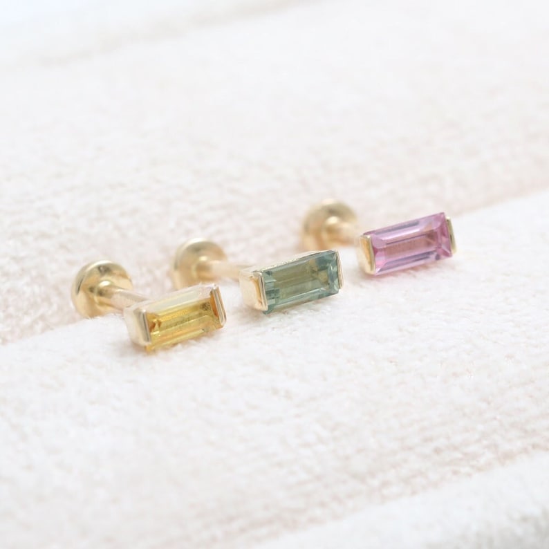 May include: Three gold stud earrings with rectangular, coloured gemstones. The gemstones are yellow, green, and pink. The earrings are displayed on a white, textured surface.