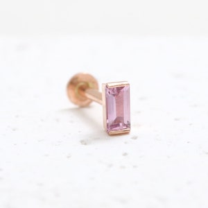 May include: A single, rose gold earring featuring a rectangular, pale pink gemstone. The earring has a post and a small, round back. The gemstone is set in a matching rose gold bezel.