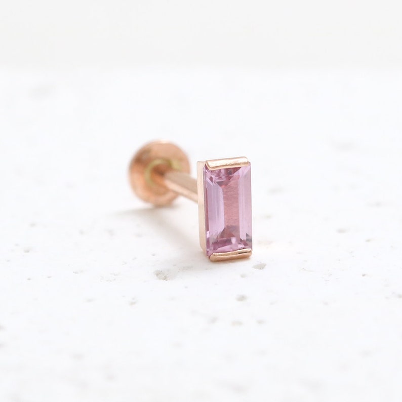 May include: A single, rose gold earring featuring a rectangular, pale pink gemstone. The earring has a post and a small, round back. The gemstone is set in a matching rose gold bezel.
