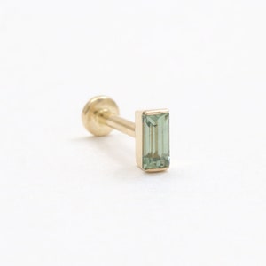 May include: A gold stud earring featuring a rectangular, pale green gemstone. The earring has a flat, round back and a slender post. The gemstone is set in a gold bezel. The earring is approximately 0.5 cm in length.