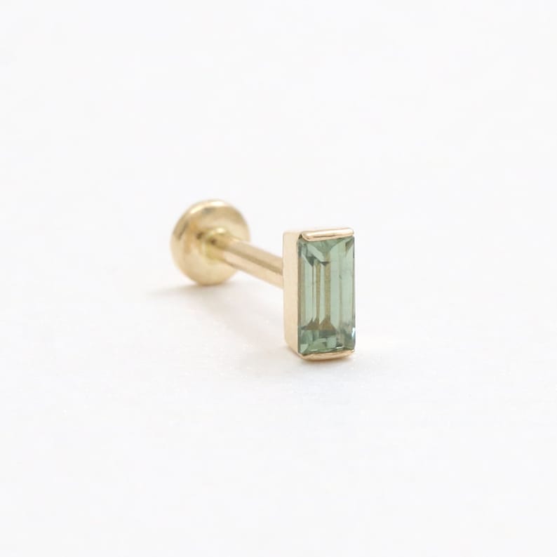 May include: A gold stud earring featuring a rectangular, pale green gemstone. The earring has a flat, round back and a slender post. The gemstone is set in a gold bezel. The earring is approximately 0.5 cm in length.