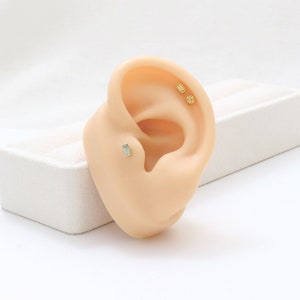 May include: A prosthetic ear displaying three pieces of jewellery. One is a rectangular, pale green stud, and the other two are small, round, gold-coloured earrings. The ear is set against a white background.