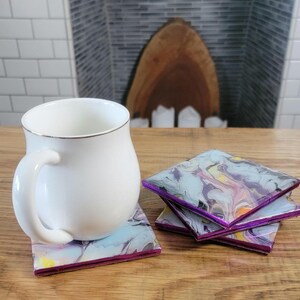 May include: A white ceramic mug with a silver rim sits on a square coaster with a swirling, abstract design in shades of purple, blue, and yellow. Three additional coasters with the same design are stacked nearby. The coasters have a purple border.