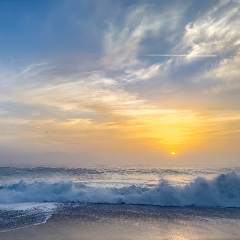May include: A scenic ocean view at sunset. The sky transitions from blue to golden yellow, with wispy clouds. Waves crash on the shore, creating white foam. The sun is a bright orb near the horizon, casting a warm glow over the water.