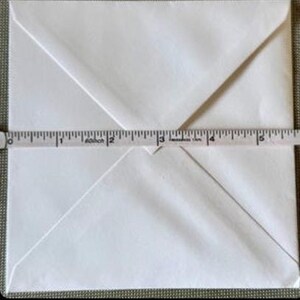 May include: A white envelope with a folded flap, measured with a tape measure. The envelope is approximately 5 inches wide. The envelope is on a neutral background.